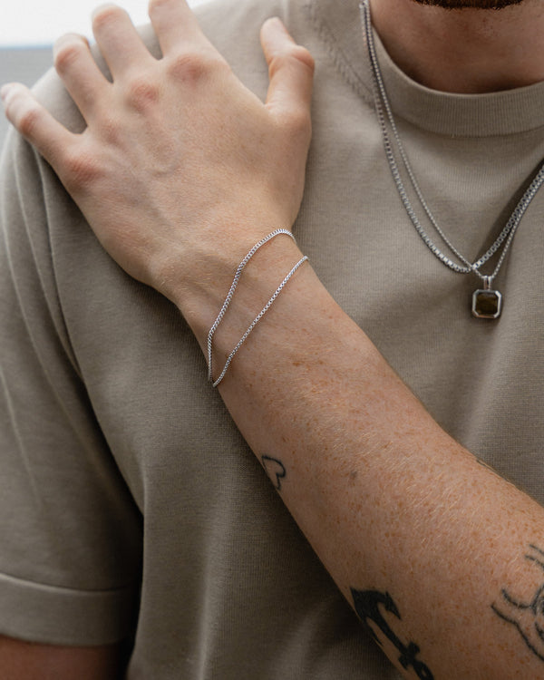 Ein Mann traegt zwei Armbänder Box Thin und Small Curb aus 925 Silber gefertigt in feiner Handarbeit das Licht spiegelt auf den glaenzenden Kettengliedern Sprezzi Fashion