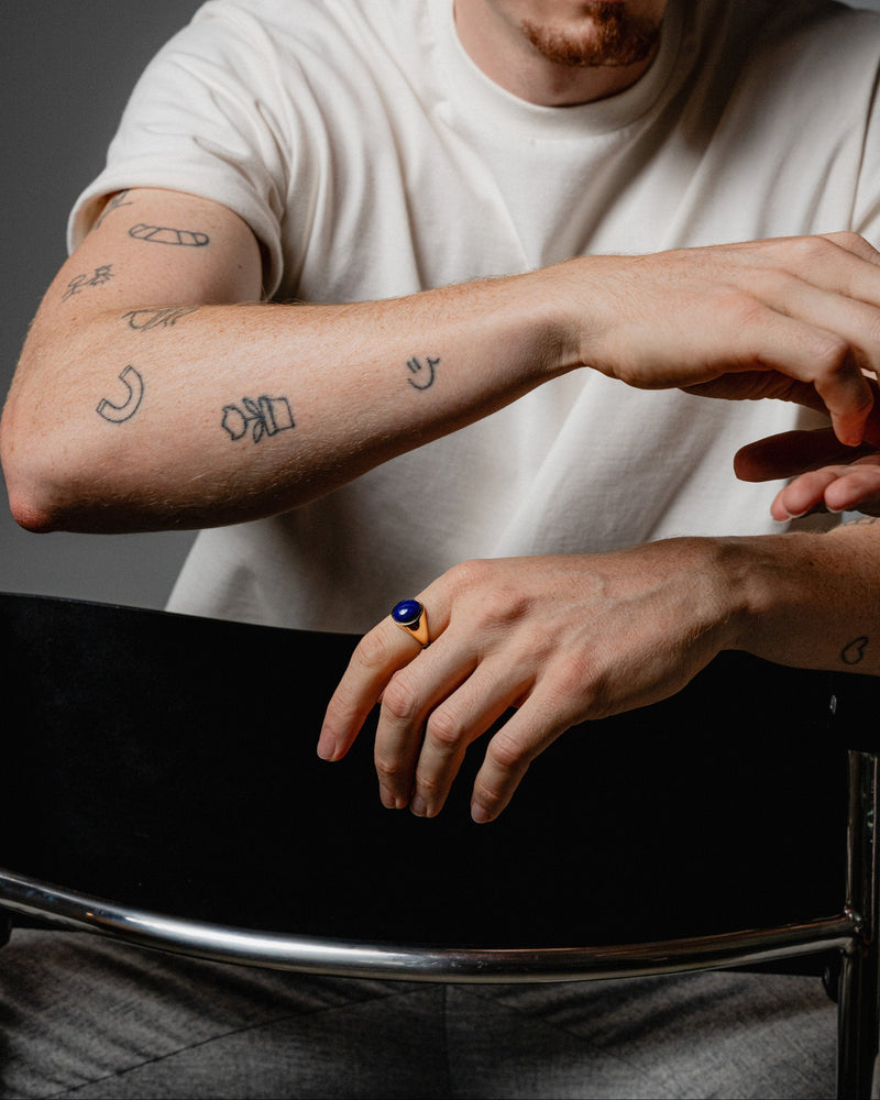 Ein Mann trägt den ovalen Lapislazuli-Siegelring aus vergoldetem 925 Sterling Silber. Der tiefblaue Stein und das goldene Finish verleihen dem Schmuck zeitlose Eleganz und luxuriösen Charakter Sprezzi Fashion
