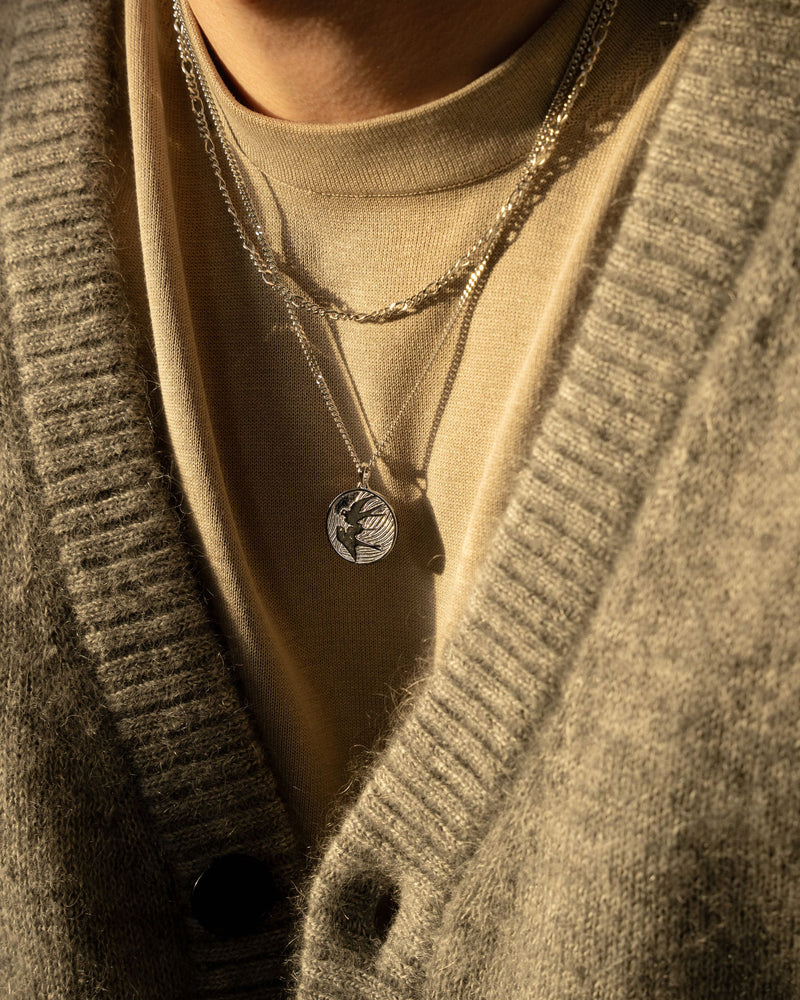 Ein Mann traegt die Peace Dove Halskette mit silberner Taube und eine Figaro Kette aus 925 Silber die Kombination aus Symbolik und Struktur steht fuer modernen Stil Sprezzi Fashion