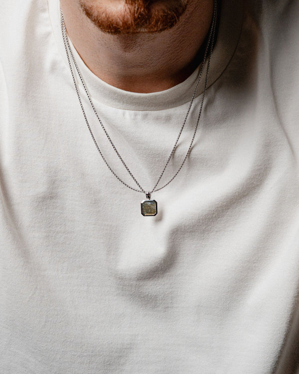 Zwei Ketten aus massivem 925 Silber liegen am Hals eines Mannes der schimmernde Larvikit Stein trifft auf kantige Box Glieder fuer einen modernen Herren Look Sprezzi Fashion