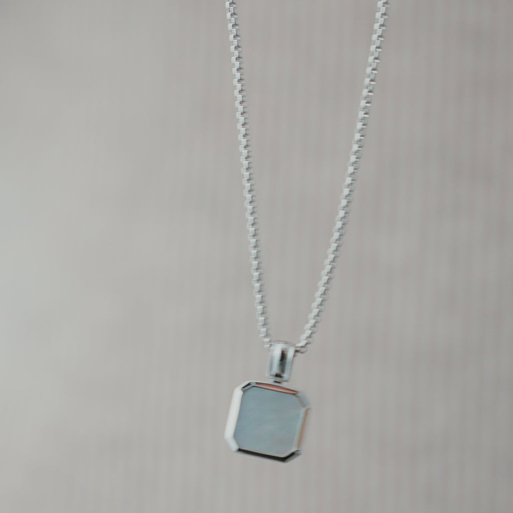 Nahaufnahme einer Herren Halskette aus 925 Sterling Silber mit eckigem weiß schimmerndem Perlmutt Anhänger, dessen Oberfläche das Licht weich reflektiert und je nach Blickwinkel unterschiedliche Nuancen zeigt Sprezzi Fashion